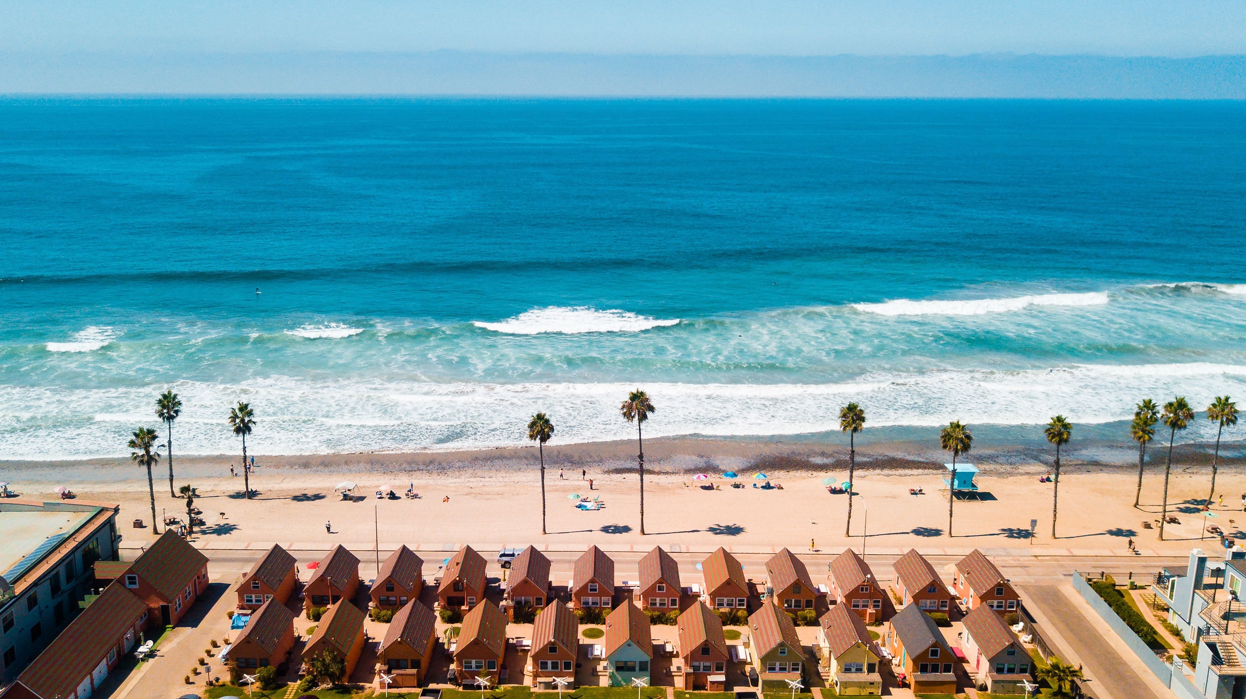 Beach cottages in a row behind the sand, palms and open ocean beyond