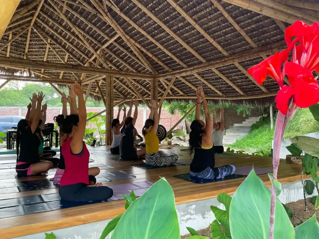A class kneeling with arms raised under the thatched roof of the shala