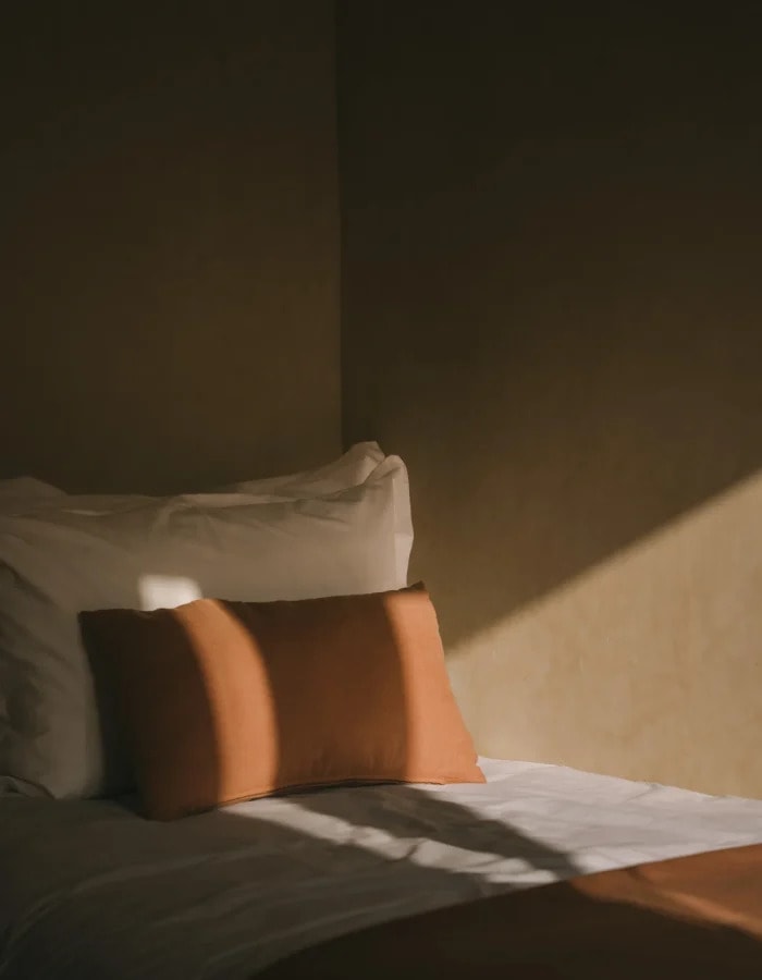 Hard afternoon light falling across a bed and a terracotta cushion